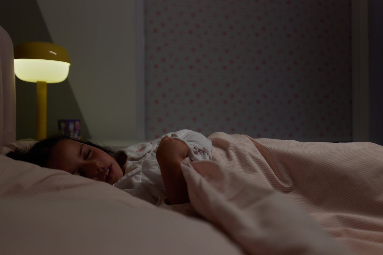 Child sleeping in pink stripped bed with a soft light on in a dimly lit room. There is a BlocOut blind fitted in the window.