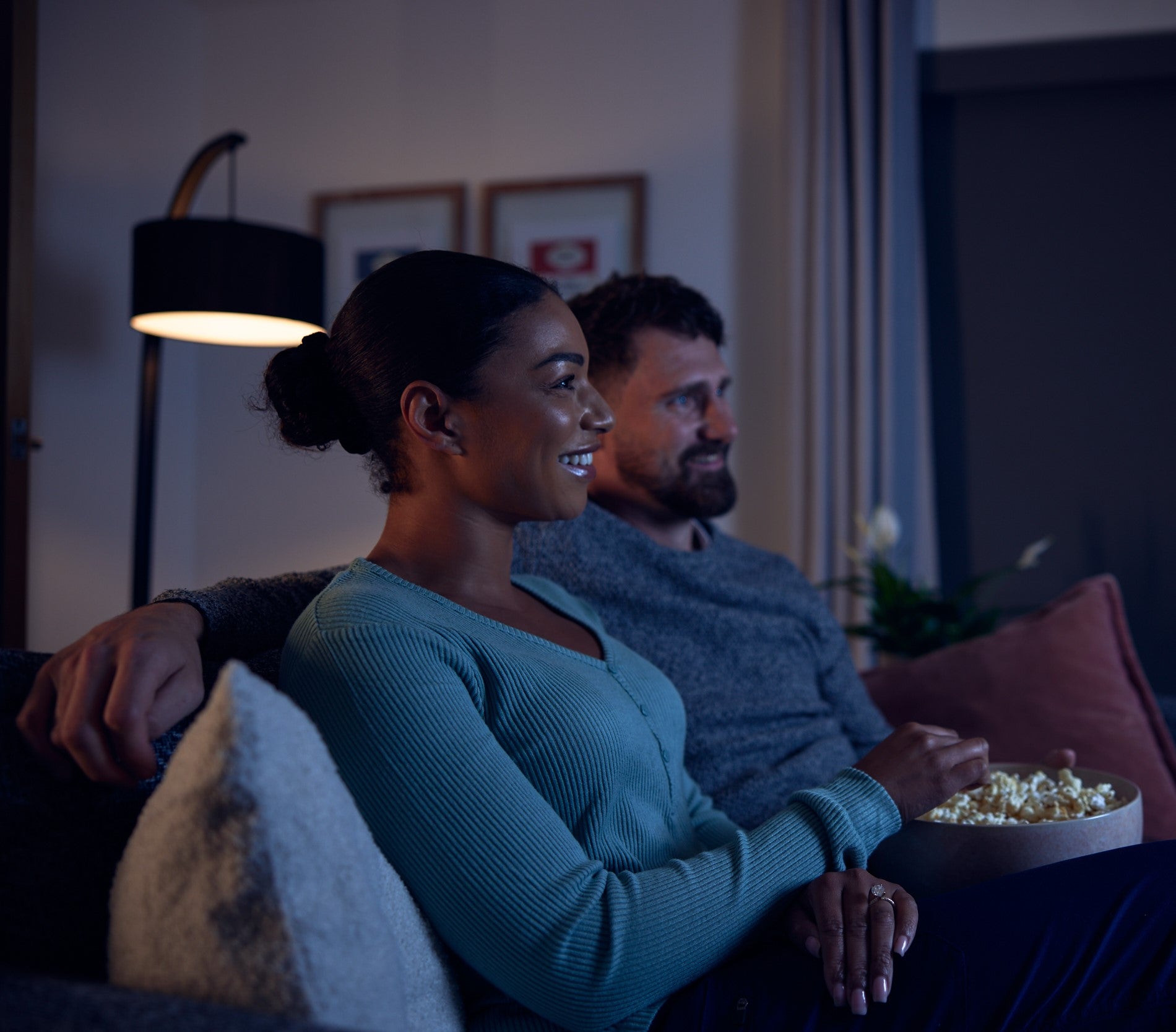 "A cozy living room scene featuring two people sitting on a couch. One is wearing a light blue sweater and holding a bowl of popcorn, while the other is dressed in a gray sweater. A stylish floor lamp with a black curved lampshade is positioned behind them, along with two framed pictures on the wall. Curtains and a green potted plant are visible in the background, enhancing the warm ambiance.