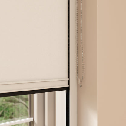 A close-up view of a window with a beige BlocOut blind partially drawn down. The shade is mounted inside the window frame, and there is a metal bead chain hanging on the right side, used to adjust the height of the shade. The wall around the window is painted in a light beige colour.