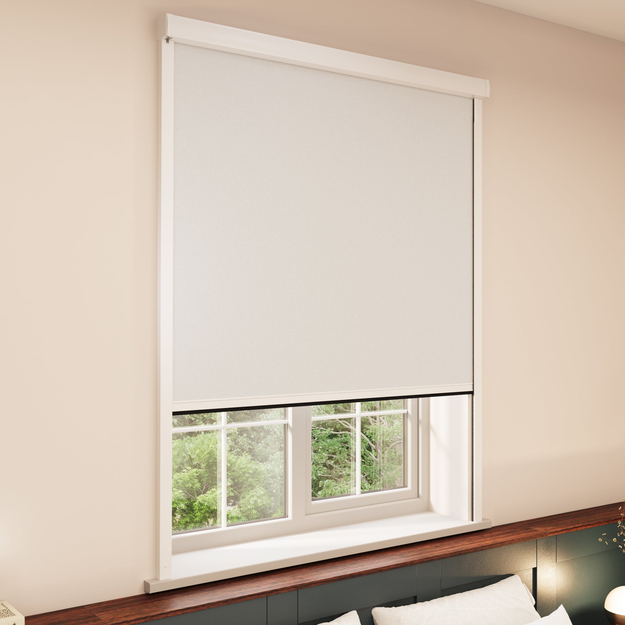 A window with a white BlocOut blind partially covering it. The window has a white frame and is set in a beige-coloured wall. Below the window, there is a wooden ledge with some pillows visible on the right side. Outside the window, green foliage can be seen.