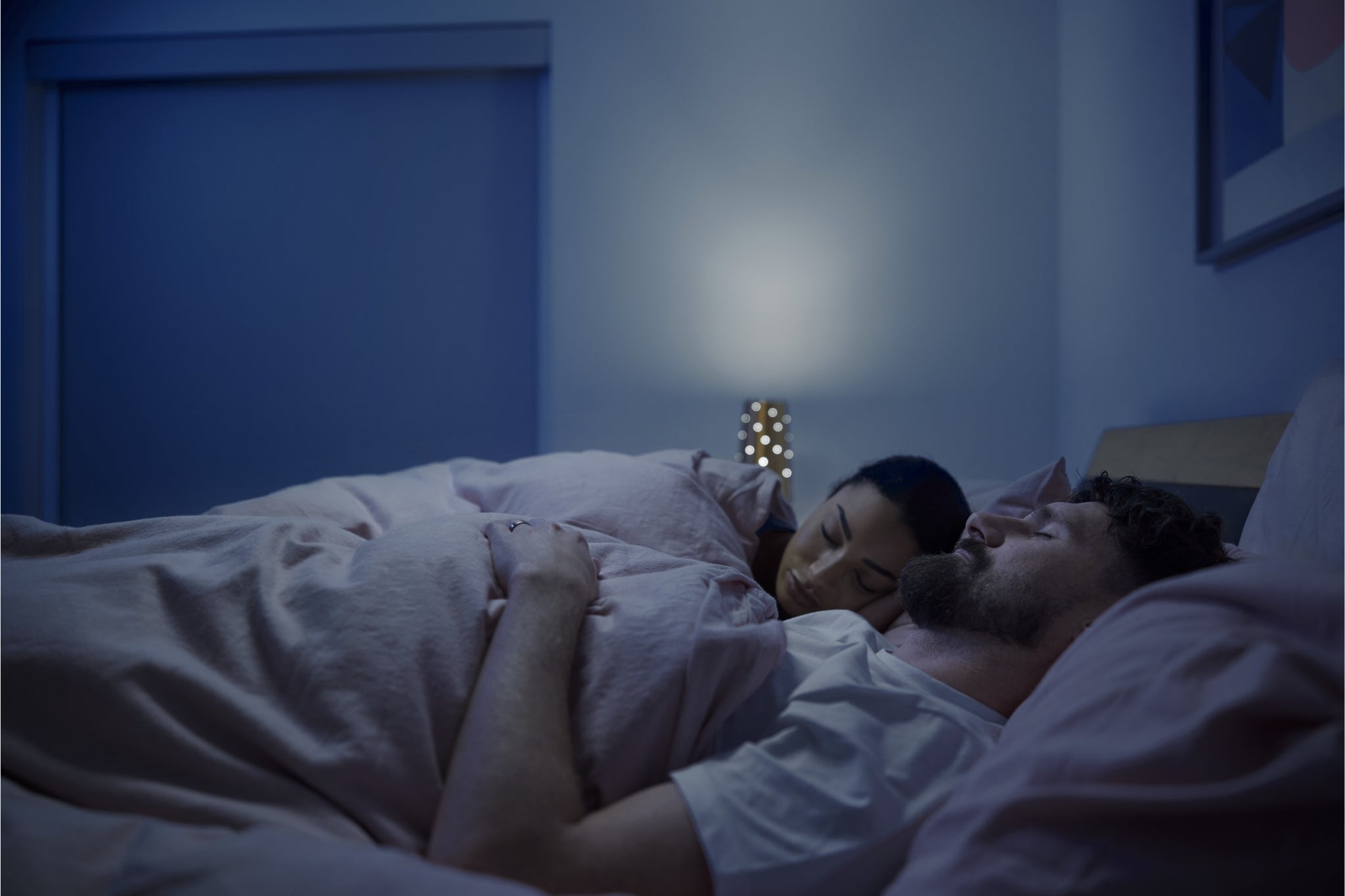 Two people lying in bed under a blanket in a dimly lit room.